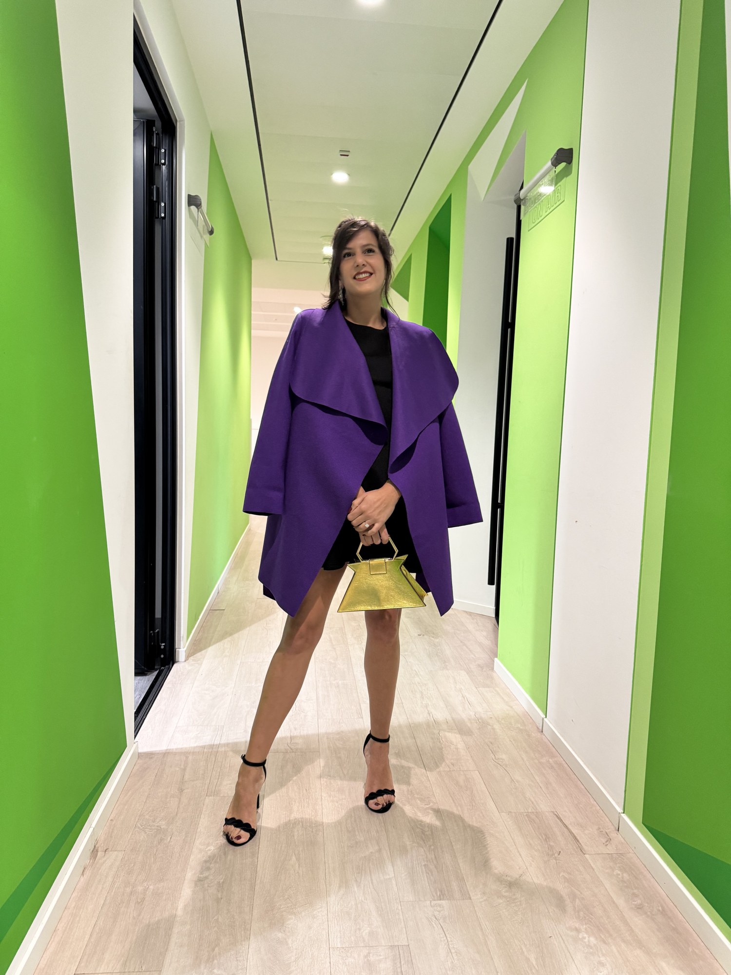 Benedetta con cappotto viola e borsa oro in un corridoio verde